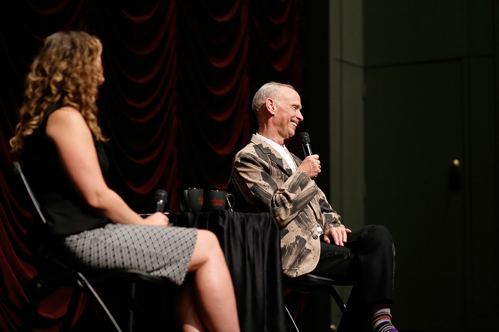 John Waters laughs while addressing the crowd while on stage with IU Cinema Associate Director Brittany Friesner.